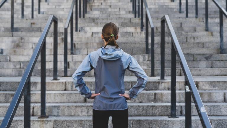 Eine Frau in grauer Sportjacke hat Müdigkeit und Energiemangel bekämpft und steht vor einer langen Treppe, bereit für ihr intensives Training, ohne dass Erschöpfung oder Müdigkeit den Sport stoppen können.