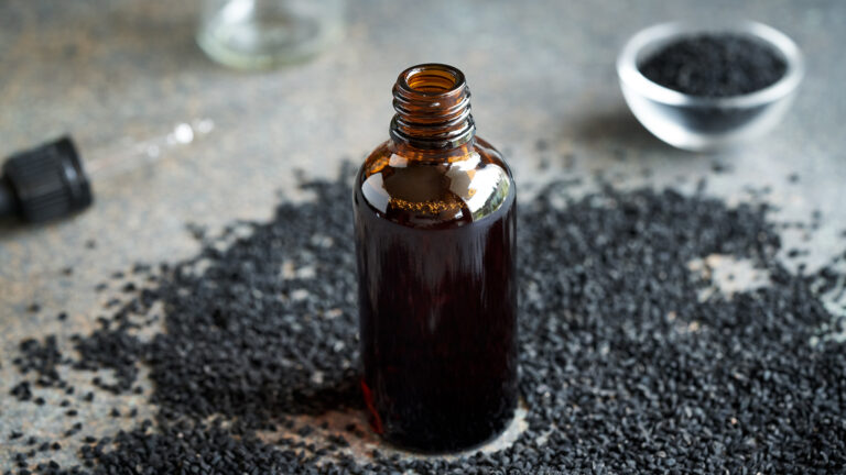 Eine geöffnete Flasche mit kaltgepresstem Schwarzkümmelöl steht umgeben von natürlichen Schwarzkümmelsamen auf einer grauen Oberfläche, mit einer Pipette und einem kleinen Schälchen voller Samen im Hintergrund.